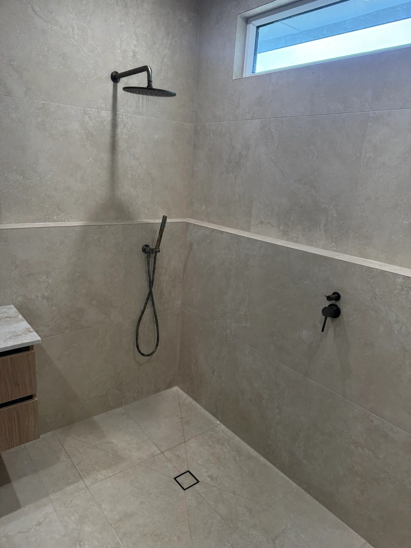 Walk-in shower with matte black wall-arm rain shower head, hand shower and mixer, travertine-look tiles and a square black floor grate